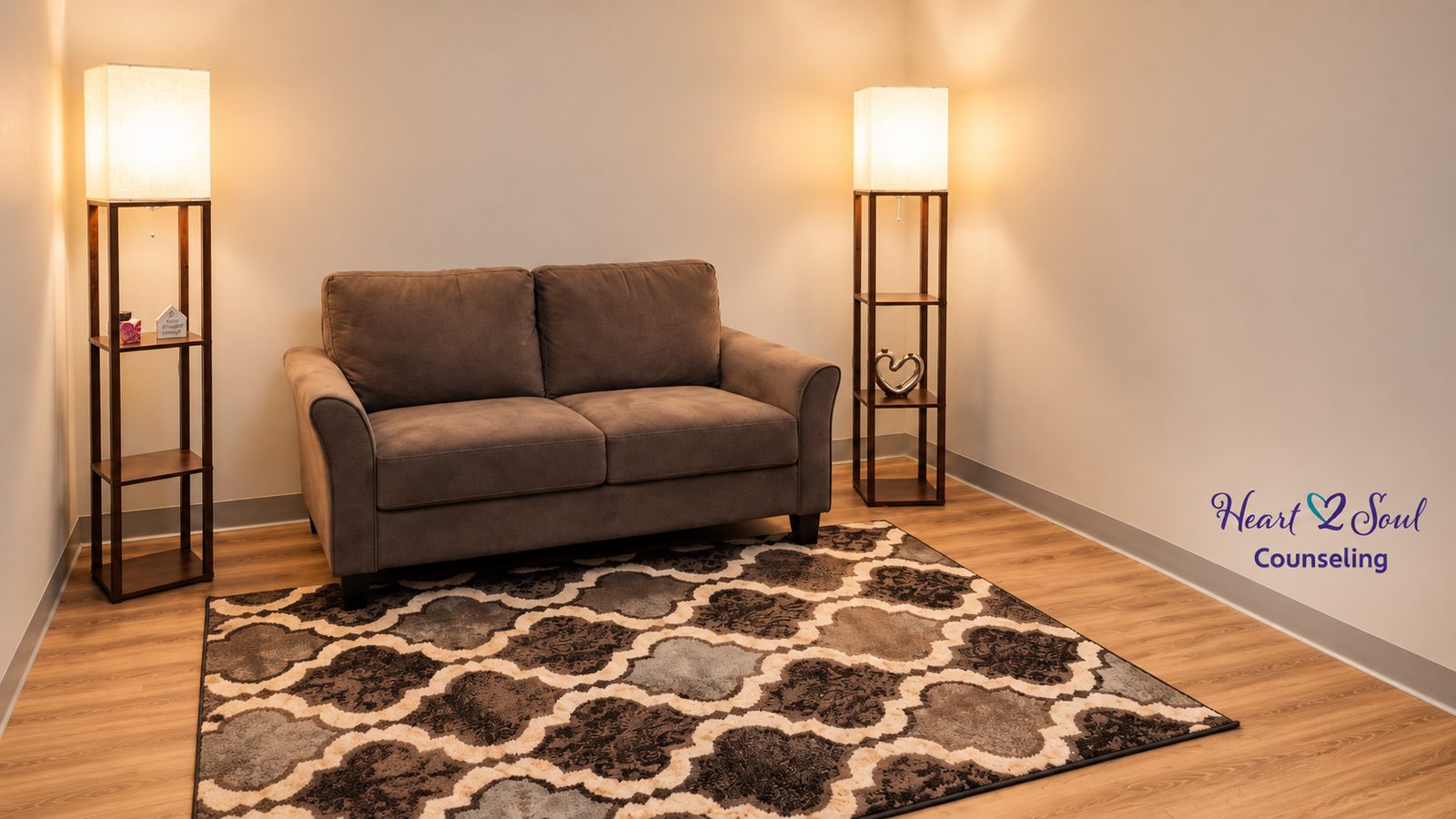 A therapy room at Heart 2 Soul Counseling with a loveseat, two shelf lamps, and the Heart 2 Soul Counseling logo on the wall